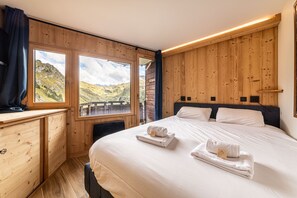 Een slaapkamer met een houten wand, een groot raam met uitzicht op de bergen, een bed met witte lakens en een nachtkastje met handdoeken.