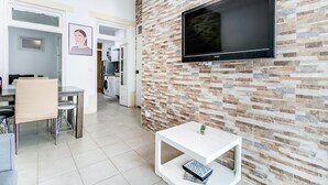 Un salon moderne avec un téléviseur à écran plat fixé à un mur dont le motif rappelle celui d’un mur de briques, une table basse blanche et un coin repas avec une chaise beige.