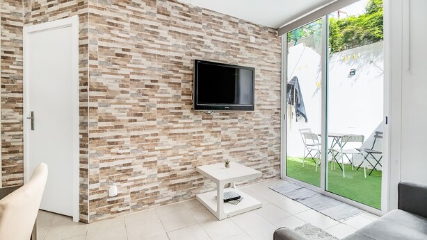 Un salon moderne avec un mur recouvert de carreaux, un téléviseur à écran plat, une table blanche et une porte-fenêtre coulissante donnant sur un espace extérieur aménagé avec des chaises.