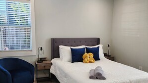 Une chambre à coucher comprenant un lit, une chaise, une table de chevet, un ventilateur de plafond et une fenêtre avec des stores.
