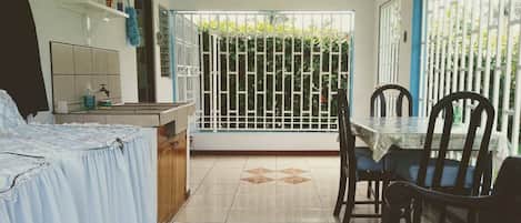 A room with a dining table, chairs, a kitchen area, and a view through a barred window.