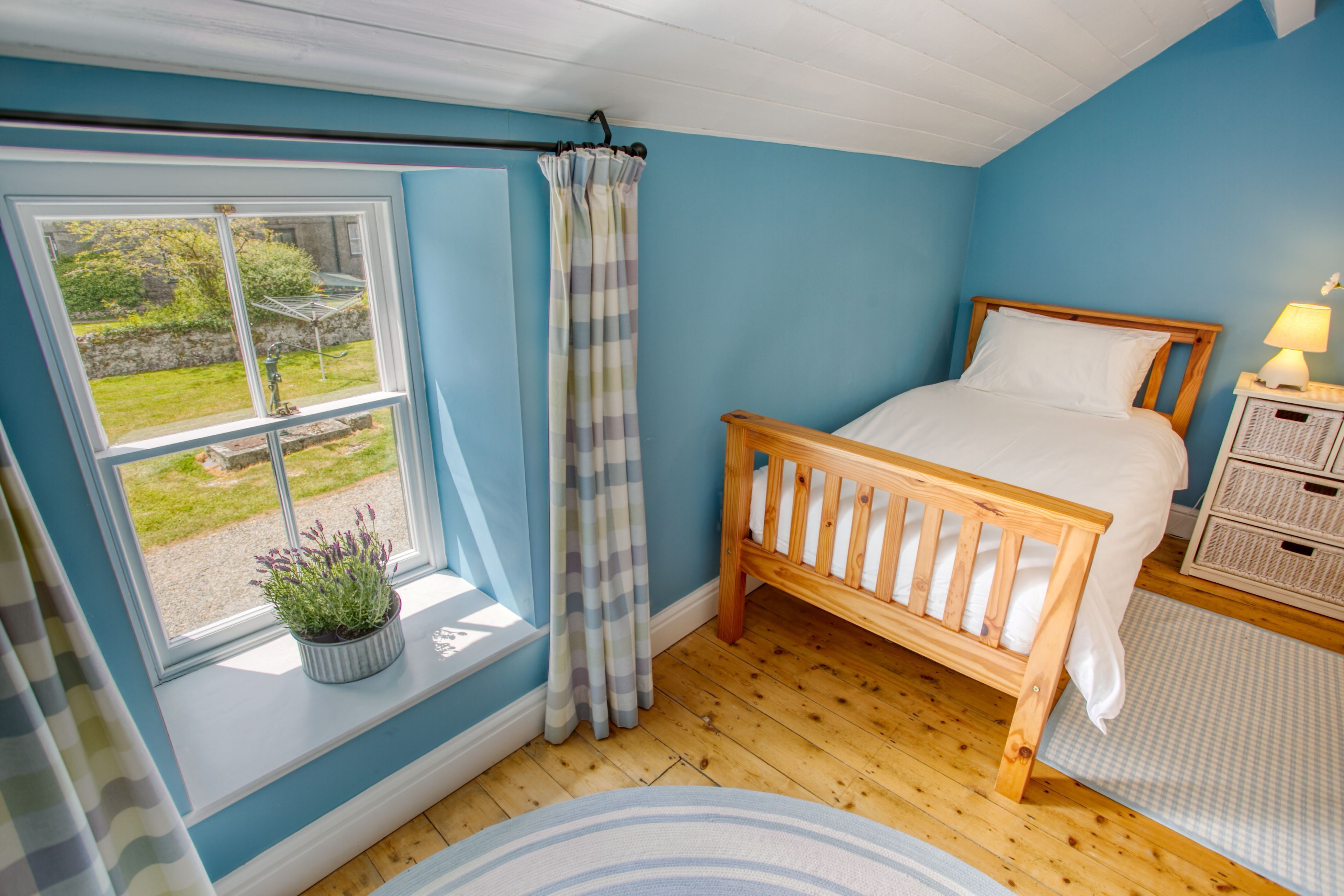 Ein Schlafzimmer mit einem hölzernen Bett, einem Fenster mit Gartenblick, einem Blumentopf auf der Fensterbank und einem Nachttisch mit Lampe.