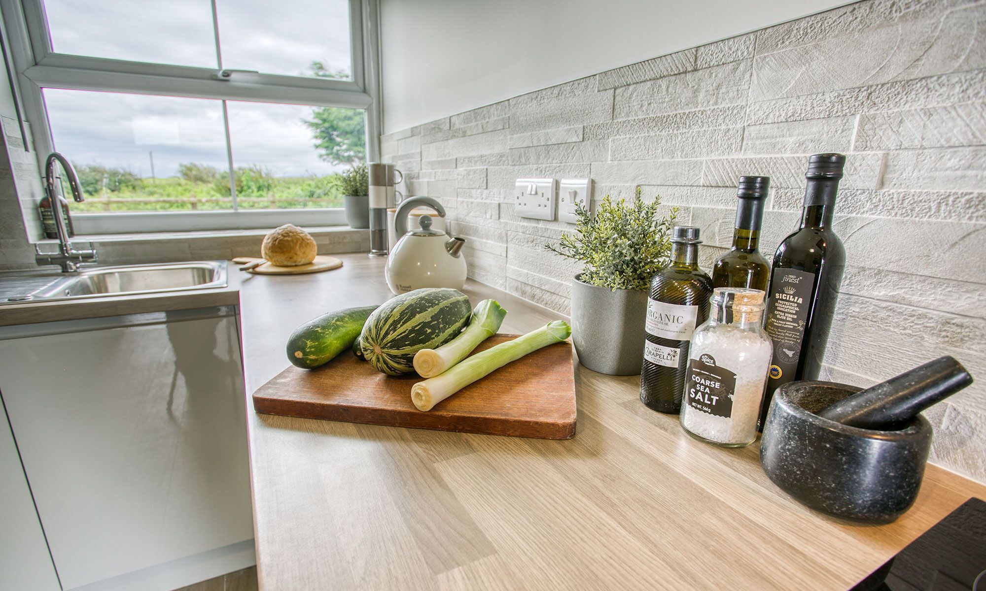 A kitchen with a wooden countertop, a white kettle, a cutting board with vegetables, a mortar and pestle, and bottles of olive oil.