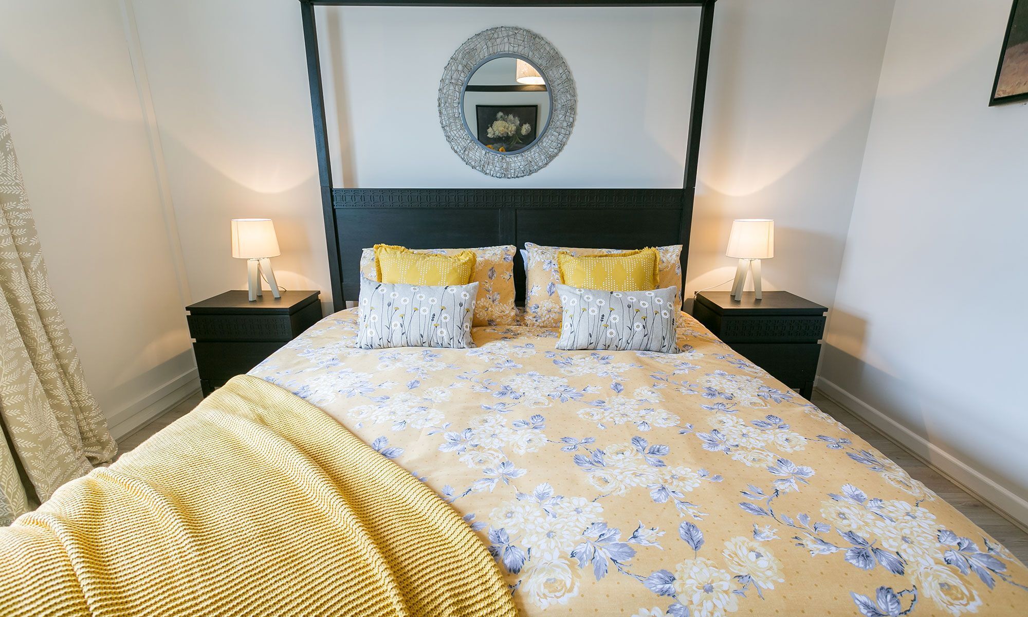 A neatly made bed with floral bedding, two bedside tables with lamps, and a round mirror above the bed.