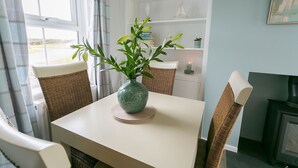 A dining area with a table, chairs, a vase with green plants, a bookshelf, and a fireplace. - Pen Y Parc Rhosneigr -  a coastal that sleeps 4 guests  in 2 bedrooms (Rhosneigr)