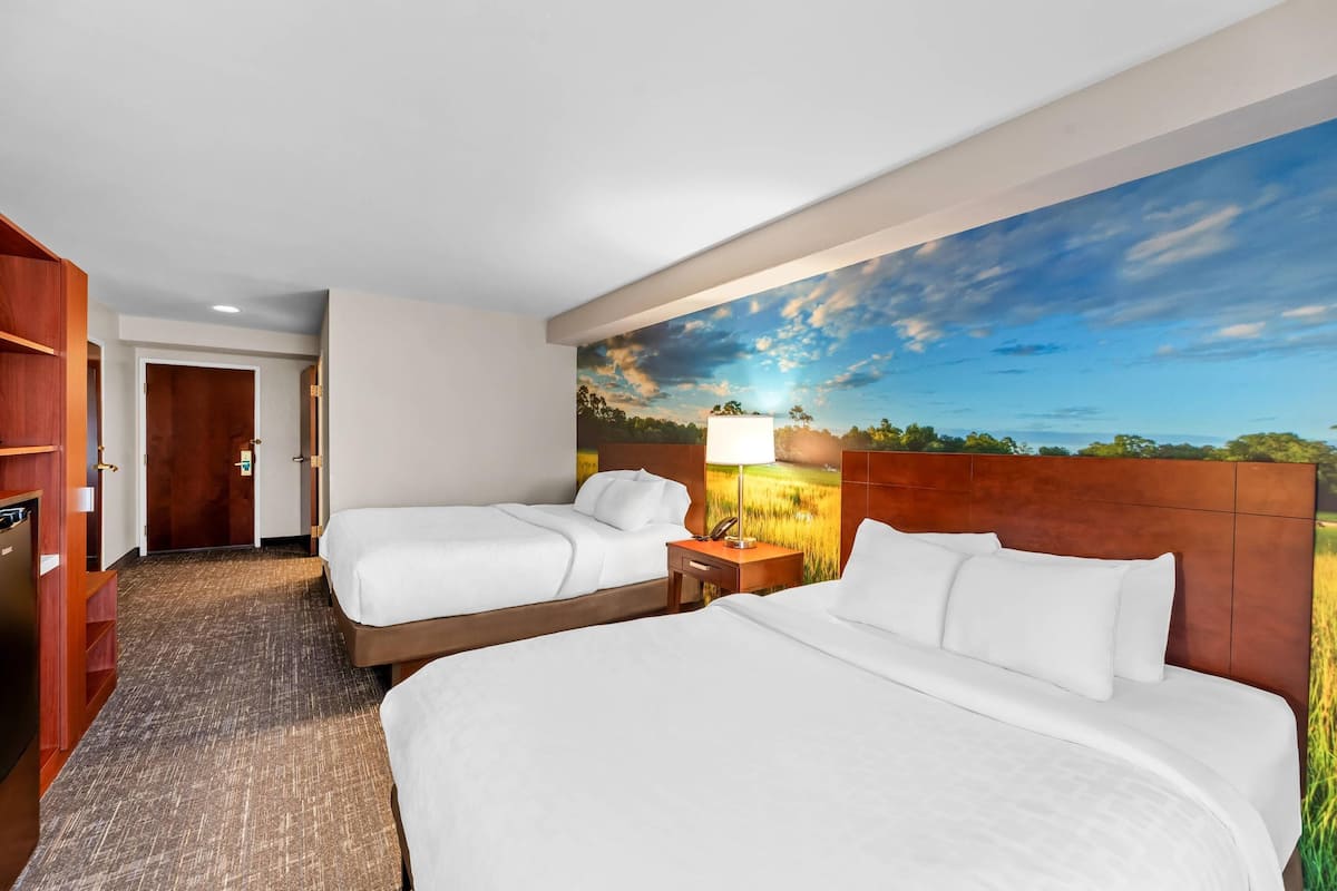 A hotel room with two beds, a wooden headboard, a nightstand with a lamp, and a large landscape mural on the wall.