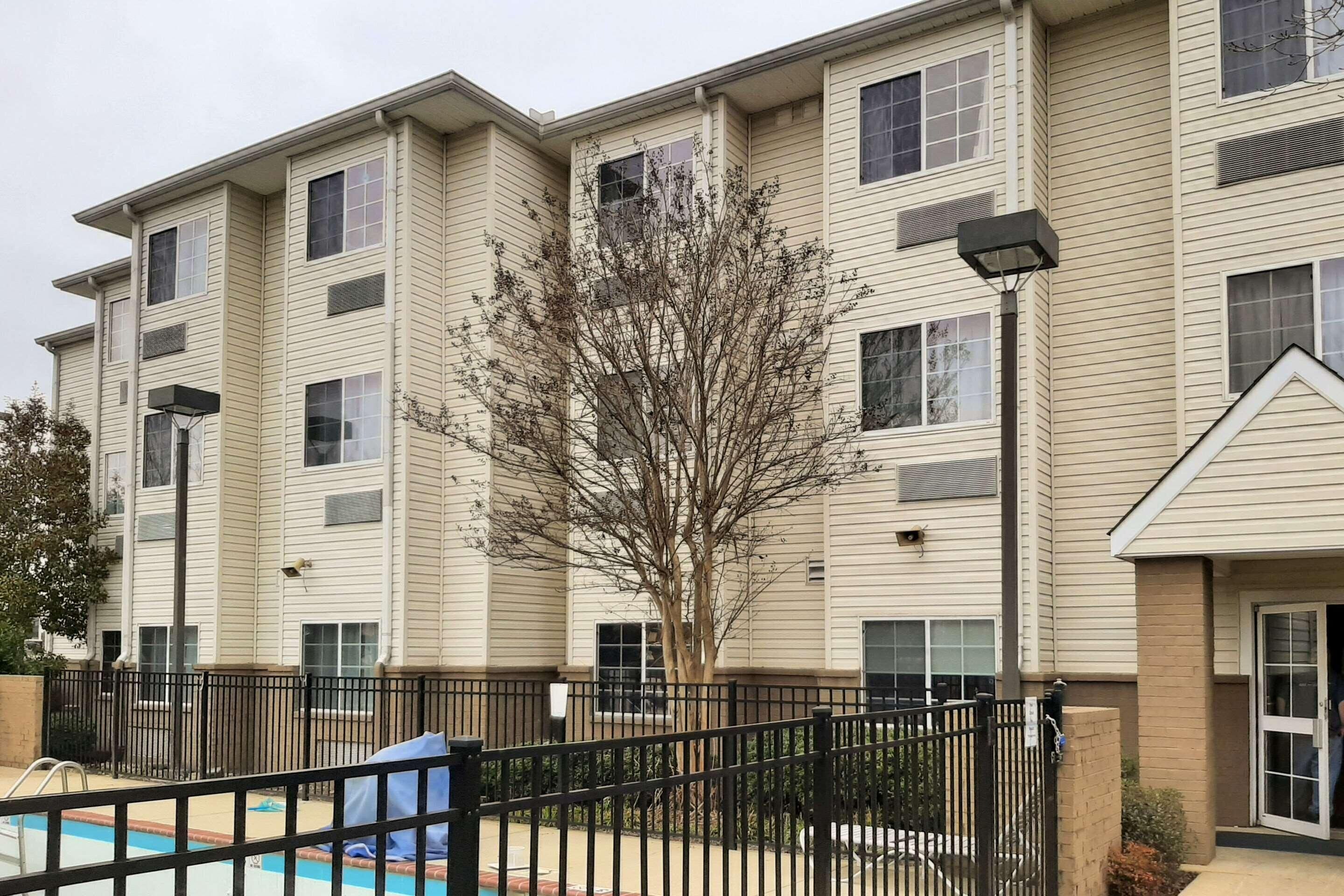 A multi-story apartment building with a swimming pool, black metal fence, and a tree in front.