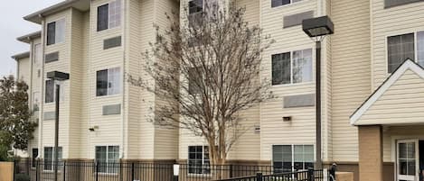 A multi-story apartment building with a swimming pool, black metal fence, and a tree in front.