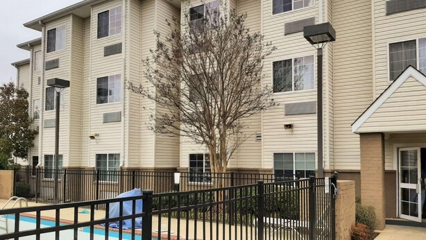 A multi-story apartment building with a swimming pool, black metal fence, and a tree in front. - Starkville Inn and Suites (Starkville)