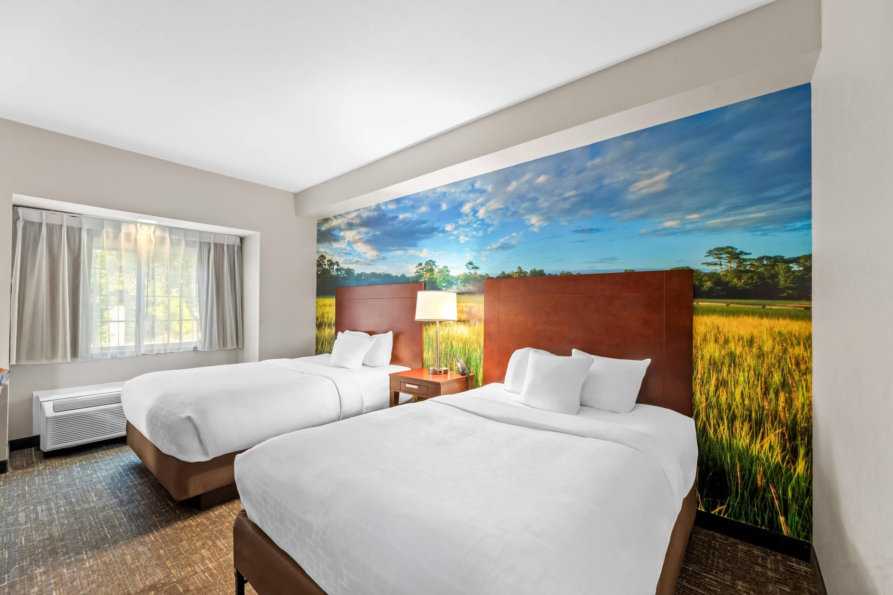 Habitación de hotel con dos camas, una ventana con cortinas, un calefactor, una mesita de noche con lámpara y un mural de un campo y cielo.