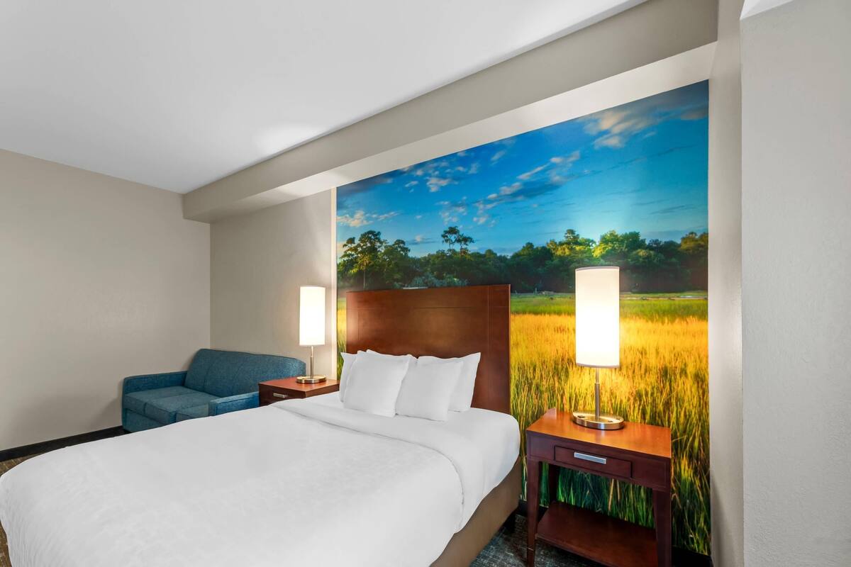 A hotel room with a large bed, a blue sofa, two bedside tables with lamps, and a wall mural of a field.
