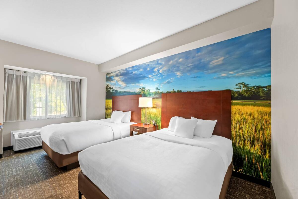A hotel room with two beds, a window with curtains, a heater, a nightstand with a lamp, and a mural of a field and sky.