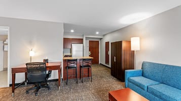 Quarto de hotel com secretária, duas cadeiras, sofá, mesa pequena, cozinha compacta e guarda-roupa.