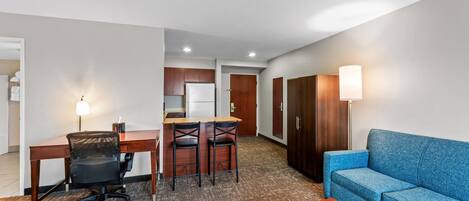 A hotel room with a desk, two chairs, a sofa, a small table, a kitchenette, and a wardrobe.