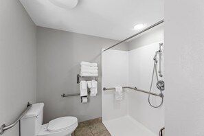 A modern bathroom with a toilet, a shower with grab bars, and a towel rack with folded towels. - Starkville Inn and Suites (Starkville)