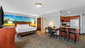 A hotel room with a bed, desk, chairs, kitchenette, and a painting on the wall. - Starkville Inn and Suites (Starkville)