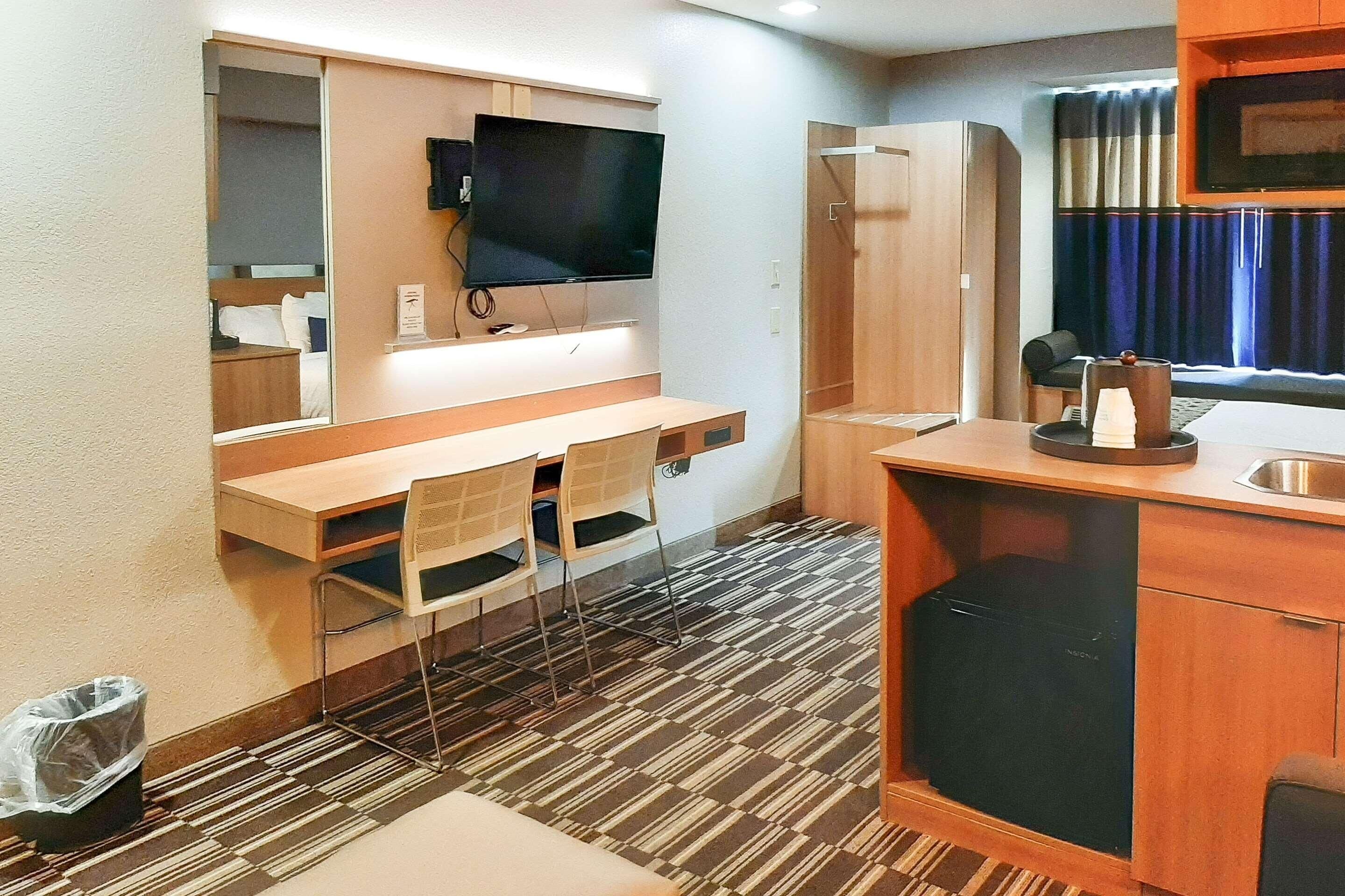 Quarto de hotel compacto com escrivaninha de madeira, duas cadeiras, televisão de tela plana na parede, mini-geladeira embutida e janela com cortinas.
