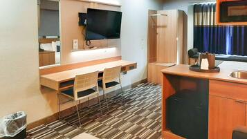 Quarto de hotel compacto com escrivaninha de madeira, duas cadeiras, televisão de tela plana na parede, mini-geladeira embutida e janela com cortinas.