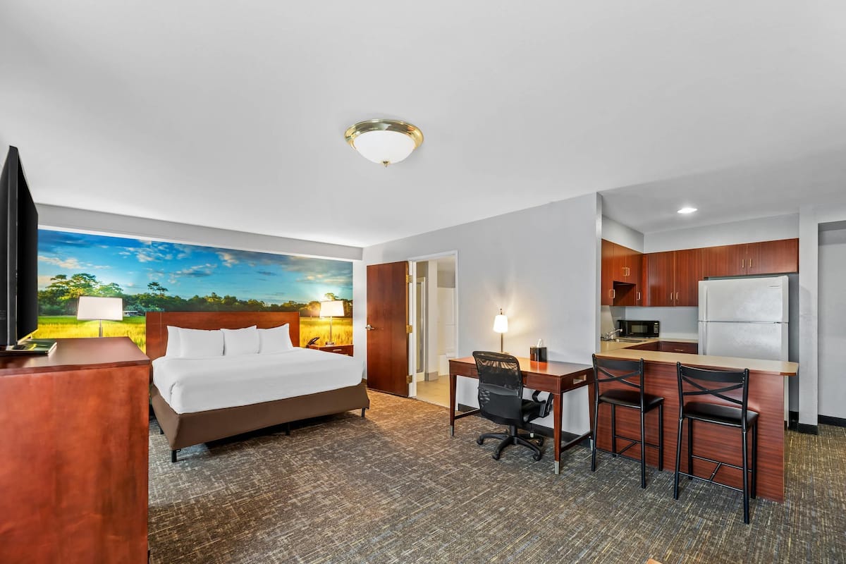 A hotel room with a bed, desk, chairs, kitchenette, and a painting on the wall.
