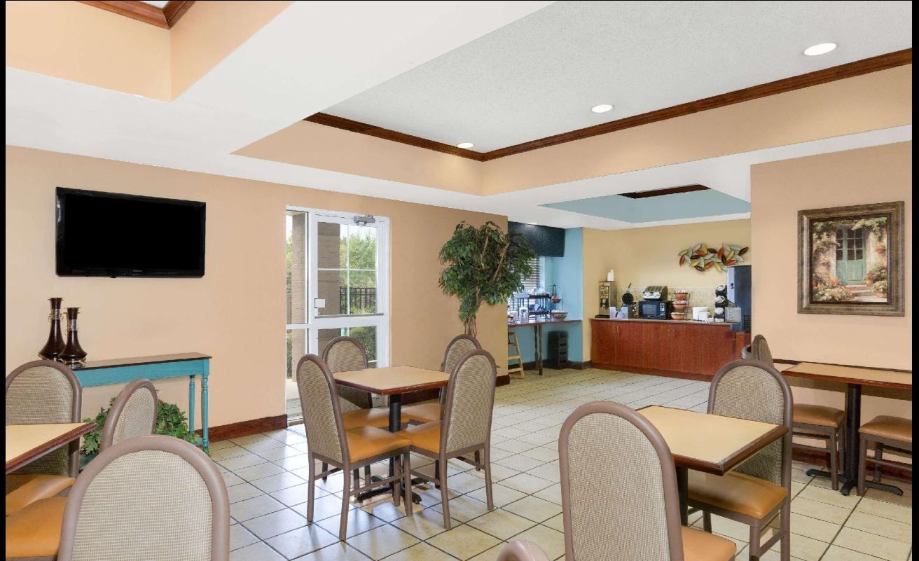 A dining area with tables and chairs, a television mounted on the wall, a plant, and a kitchen area in the background.