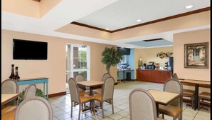 A dining area with tables and chairs, a television mounted on the wall, a plant, and a kitchen area in the background. - Starkville Inn and Suites (Starkville)