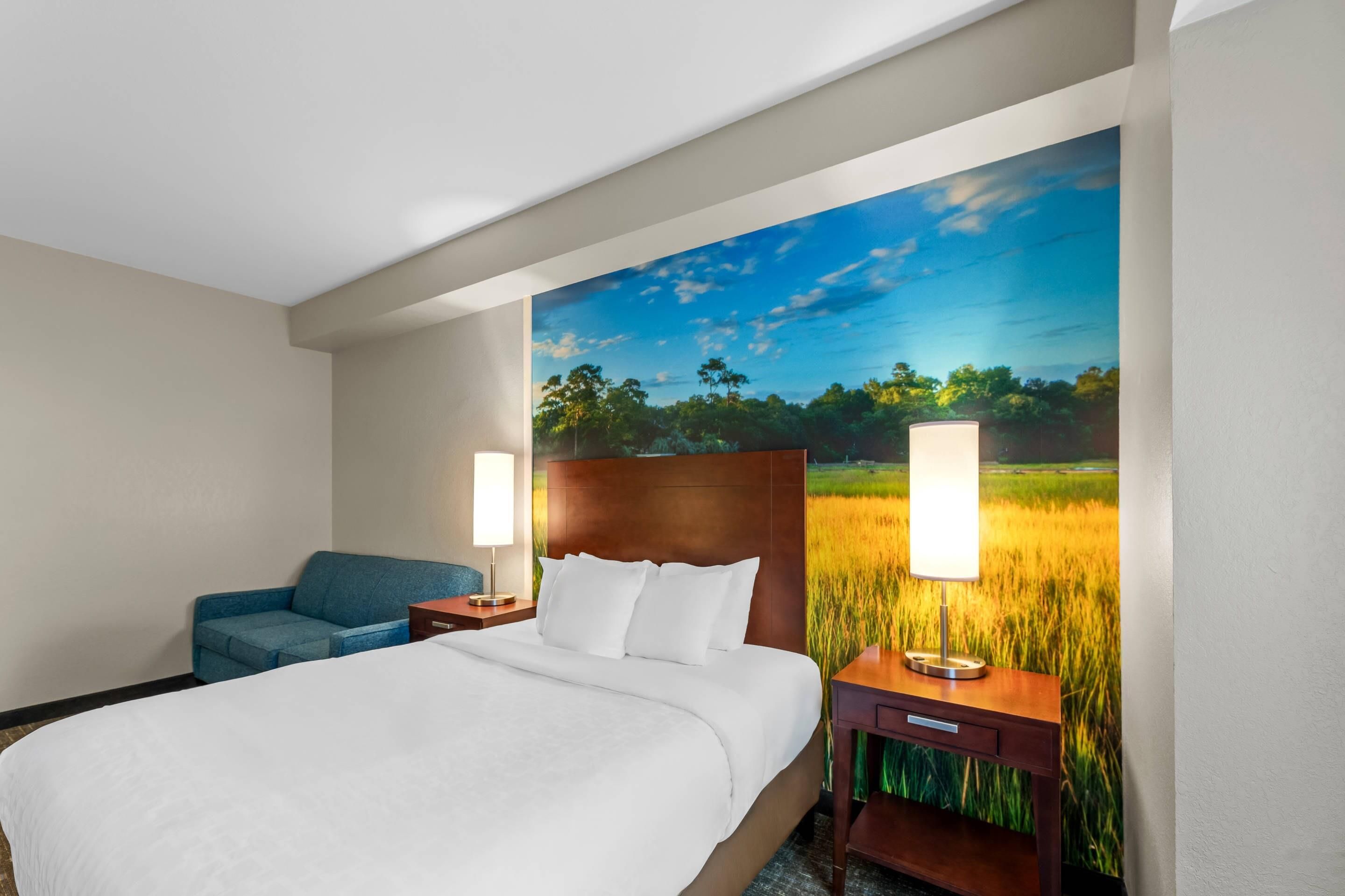 A hotel room with a large bed, a blue sofa, two bedside tables with lamps, and a wall mural of a field.