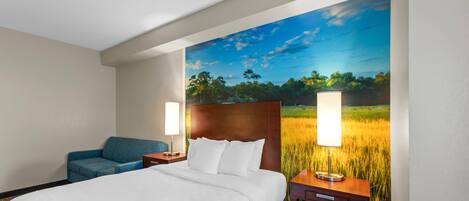 A hotel room with a large bed, a blue sofa, two bedside tables with lamps, and a wall mural of a field.
