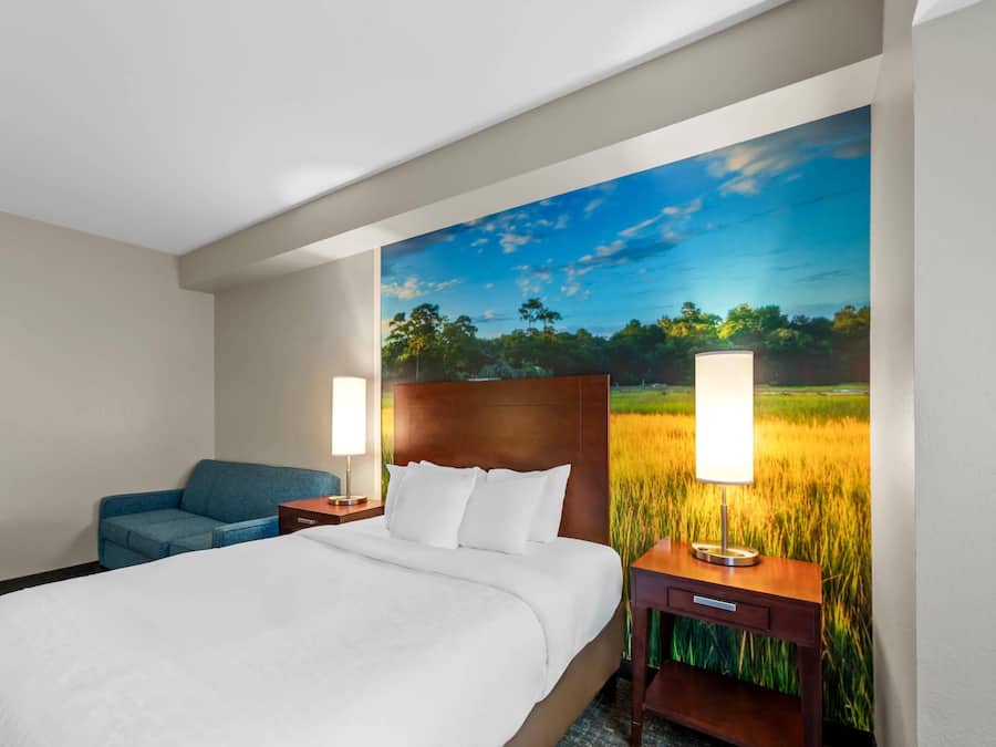 A hotel room with a large bed, a blue sofa, two bedside tables with lamps, and a wall mural of a field.