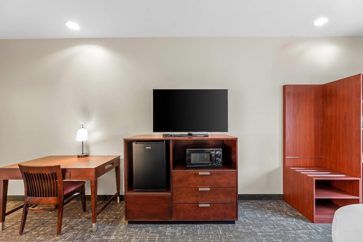 A compact room with a wooden desk, a chair, a built-in refrigerator, a microwave, a flat-screen TV, and a wardrobe.