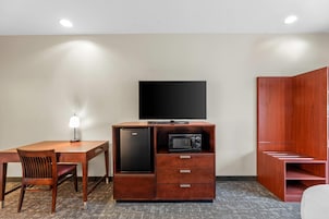 A compact room with a wooden desk, a chair, a built-in refrigerator, a microwave, a flat-screen TV, and a wardrobe.