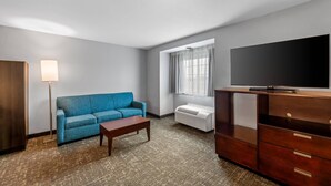 A room with a blue sofa, a wooden coffee table, a flat-screen TV on a wooden stand, a floor lamp, and a window with curtains. - Starkville Inn and Suites (Starkville)