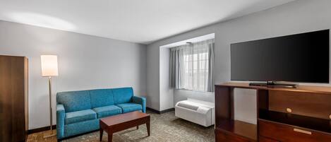 A room with a blue sofa, a wooden coffee table, a flat-screen TV on a wooden stand, a floor lamp, and a window with curtains.