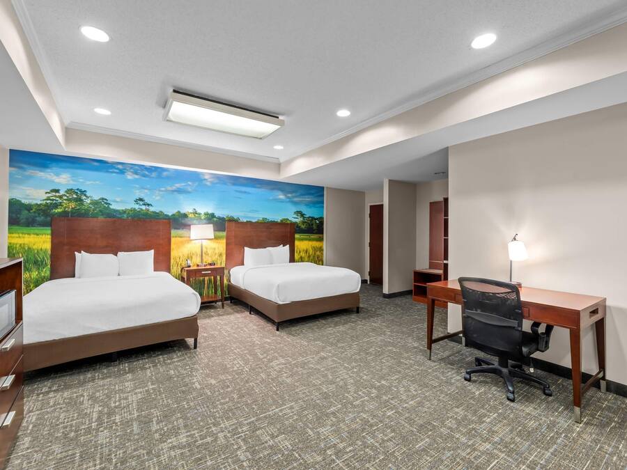 A hotel room with two beds, a desk, a chair, a TV, and a large mural of a landscape.