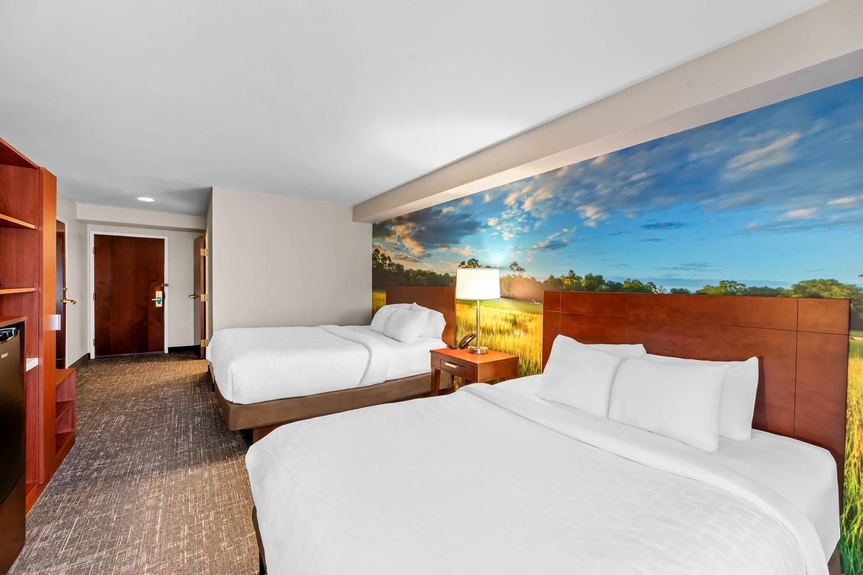 Ein Hotelzimmer mit zwei Betten, einem hölzernen Kopfteil, einem Nachttisch mit Lampe und einer großen Wandmalerei mit Landschaftsmotiv.