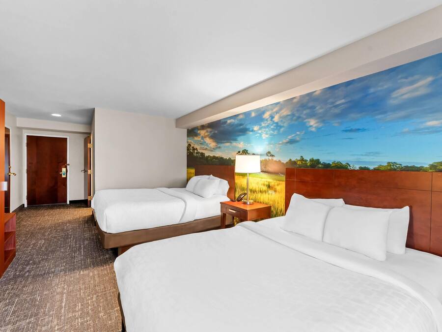 Habitación de hotel con dos camas, cabecero de madera, mesita de noche con lámpara y un mural grande con un paisaje en la pared.