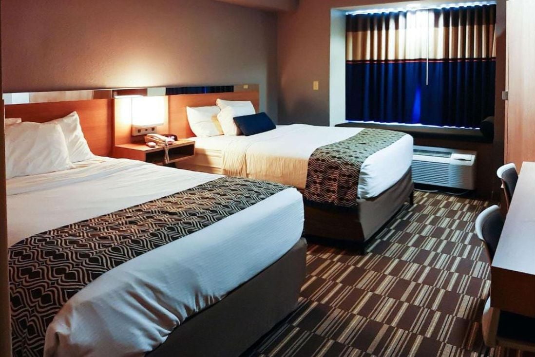 A hotel room with two beds, a desk, a chair, a window with curtains, and a wall-mounted air conditioning unit.