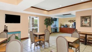 A dining area with tables and chairs, a television mounted on the wall, a plant, and a kitchen area in the background. - Starkville Inn and Suites (Starkville)