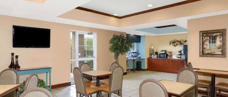 A dining area with tables and chairs, a television mounted on the wall, a plant, and a kitchen area in the background.