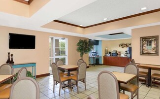A dining area with tables and chairs, a television mounted on the wall, a plant, and a kitchen area in the background.
