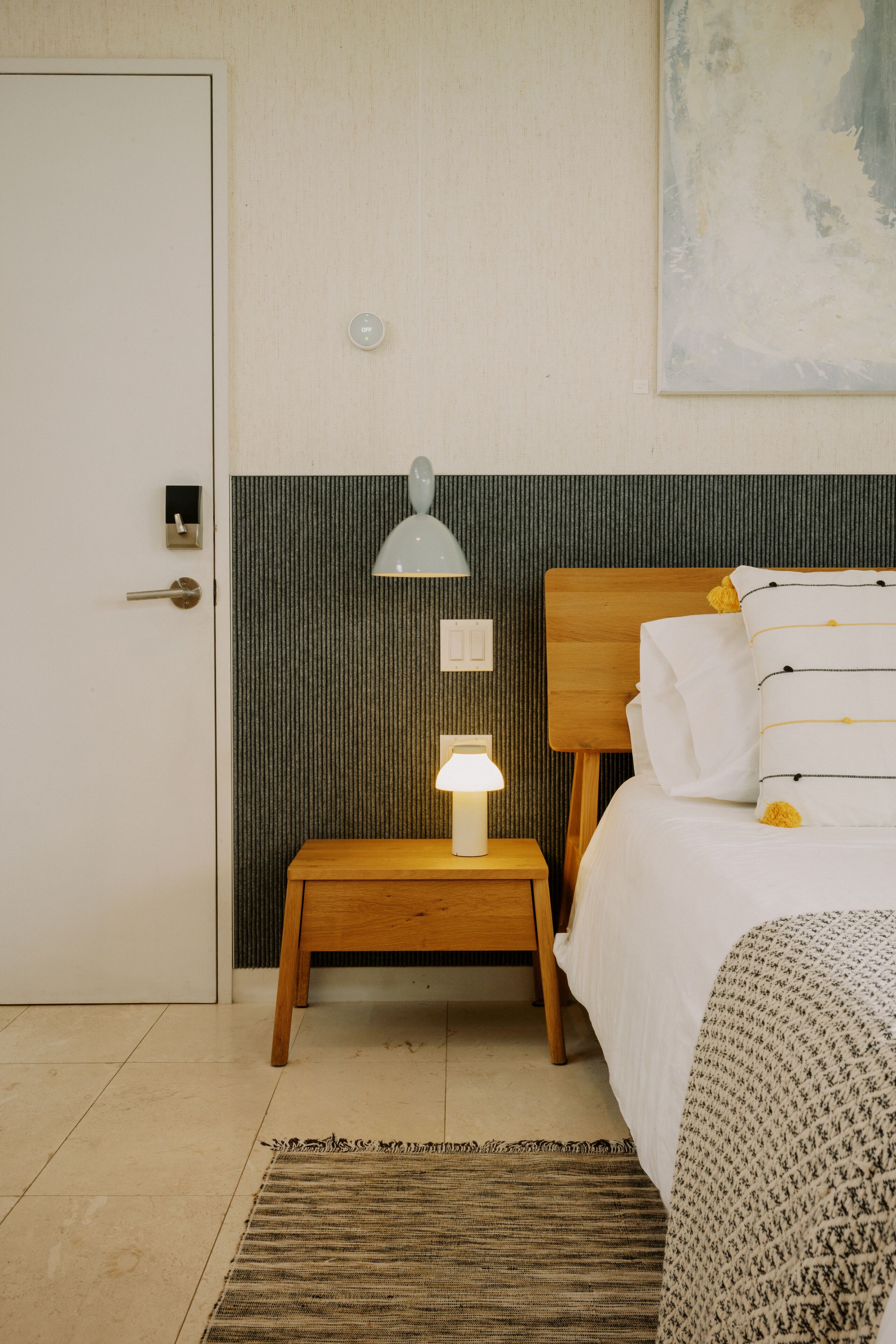 Una habitación de hotel con una cama de madera, una mesita de noche con lámpara, una puerta y una pared texturizada.