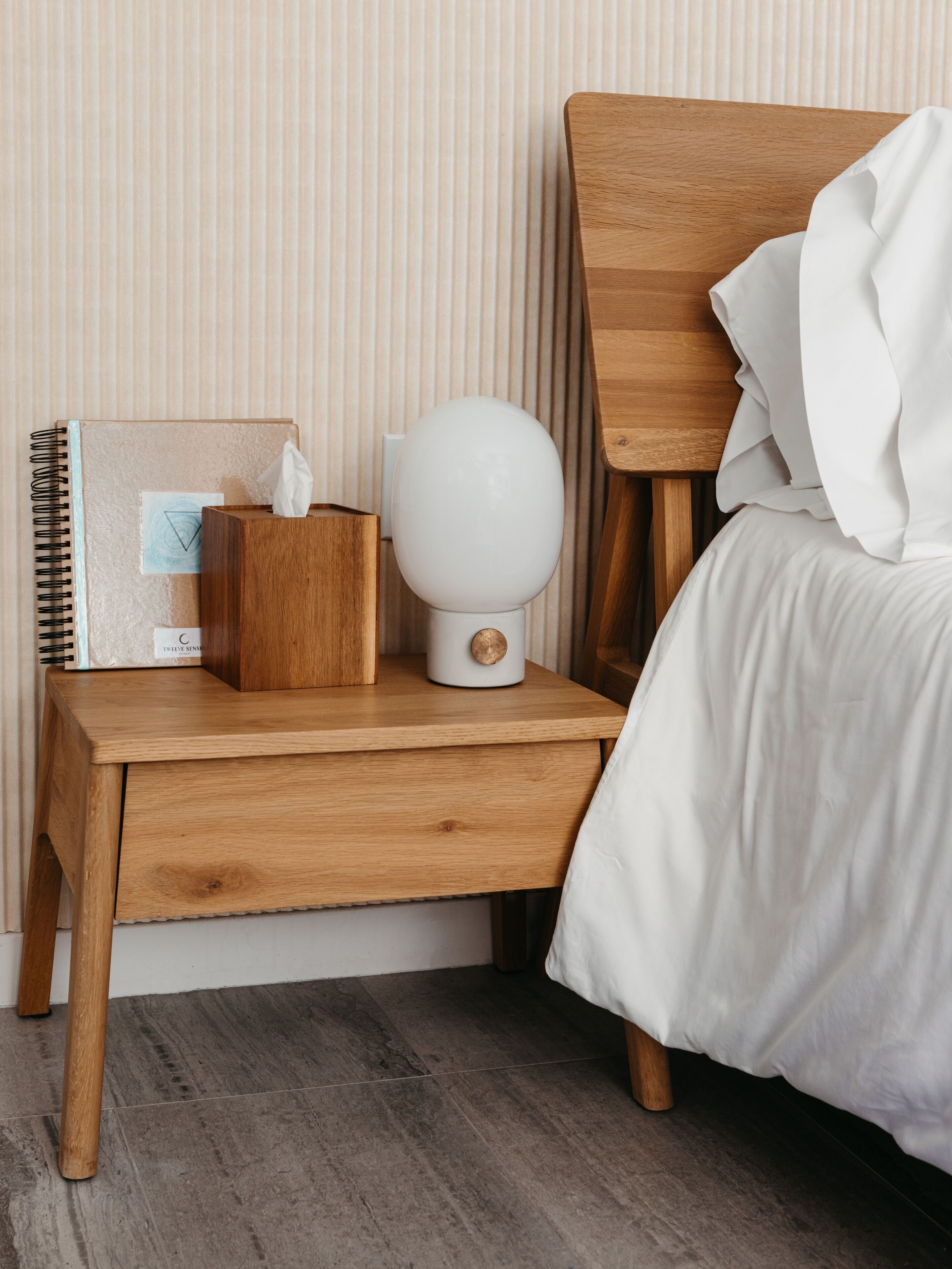 A neatly made wooden bedside table with a notebook, a tissue box, and a white lamp.