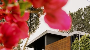 Un bâtiment moderne à deux étages, doté d’une façade en bois, d’un garage et d’un jardin bien entretenu où poussent diverses plantes et fleurs.