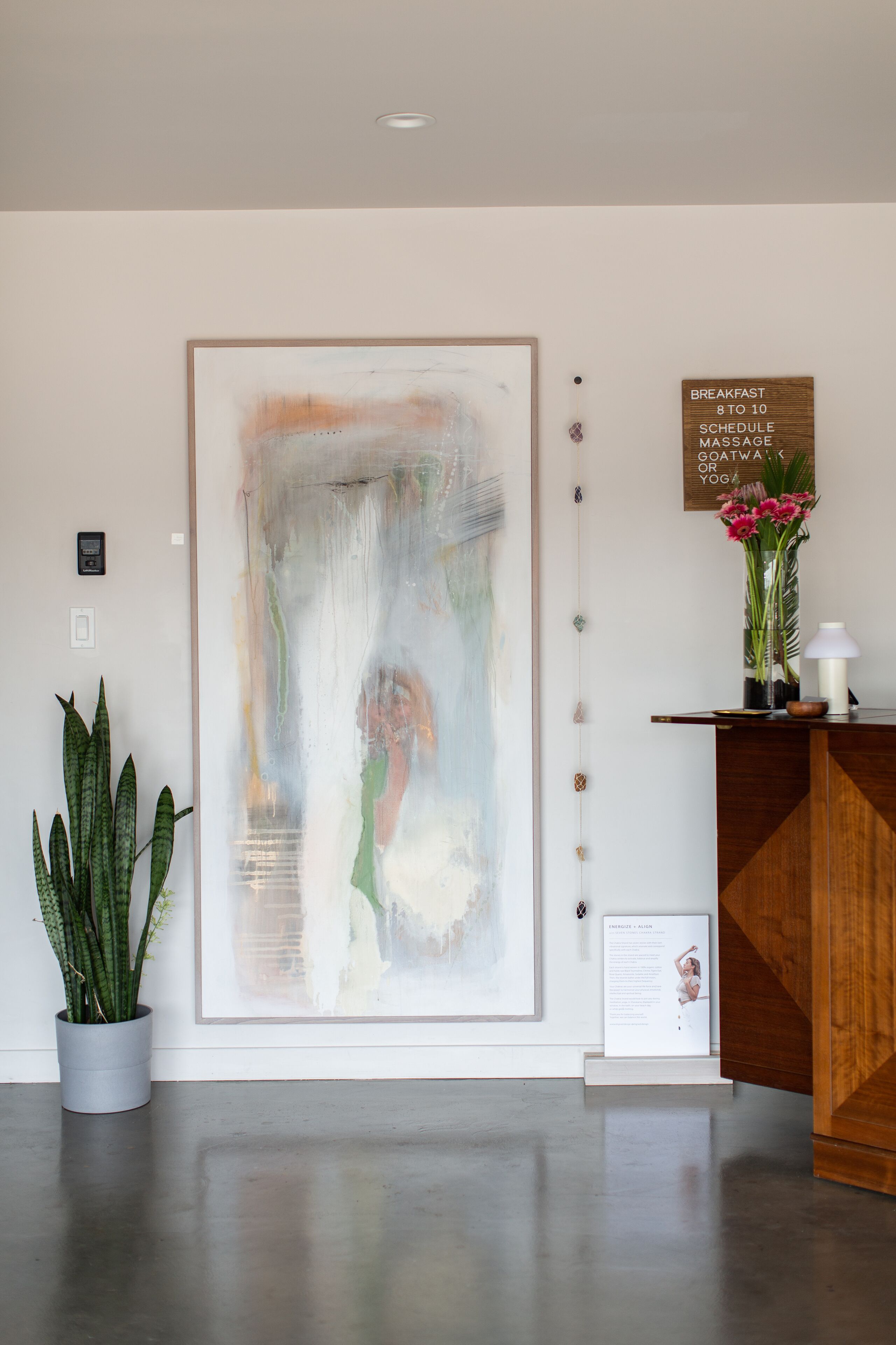 Un espace d’accueil agrémenté d’un grand tableau abstrait, d’un comptoir de réception en bois, d’une plante en pot et d’un panneau indiquant les heures du petit-déjeuner.