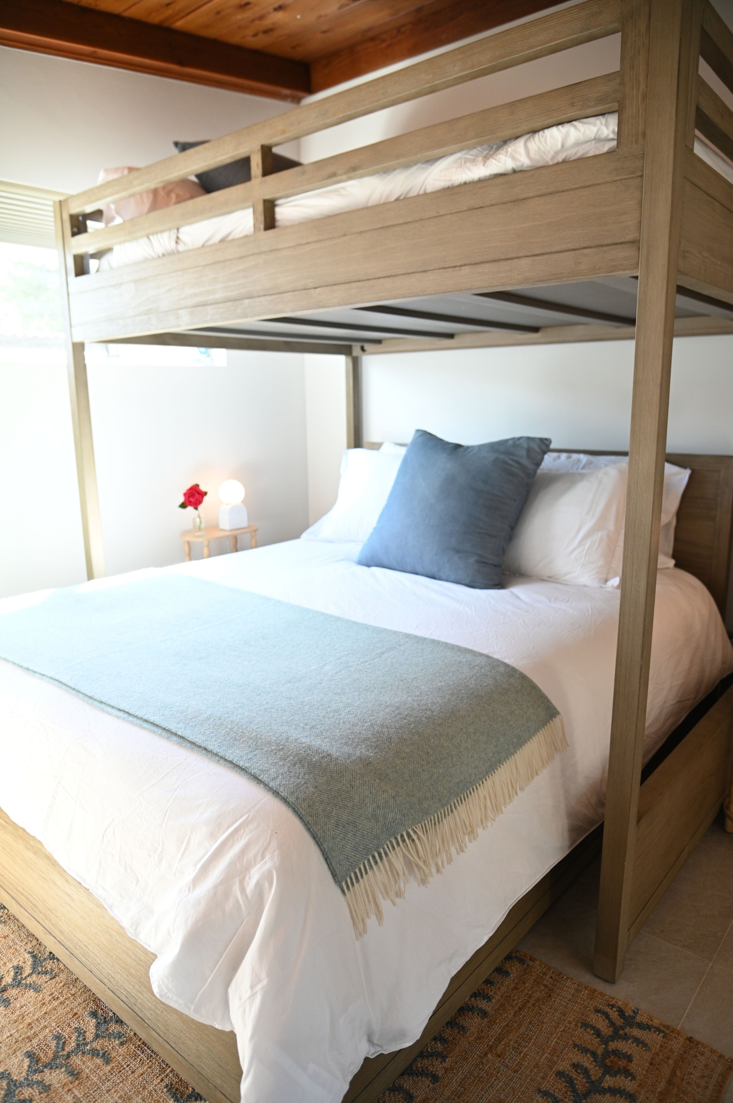 A bunk bed with a wooden frame, white bedding, a blue pillow, and a bedside table with a lamp and a vase of flowers.