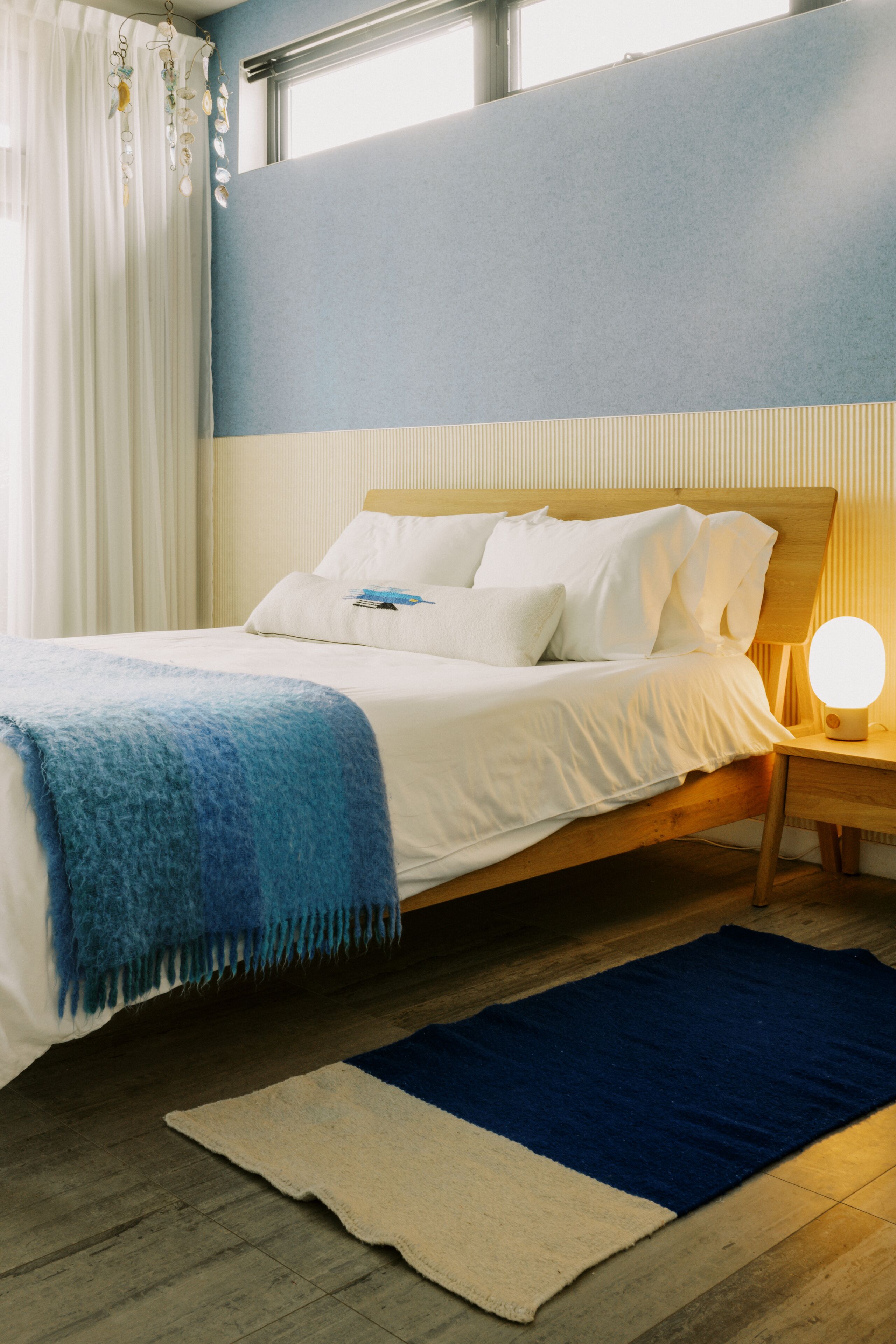 A neatly made bed with a blue blanket, a bedside lamp, and a blue rug on a wooden floor.