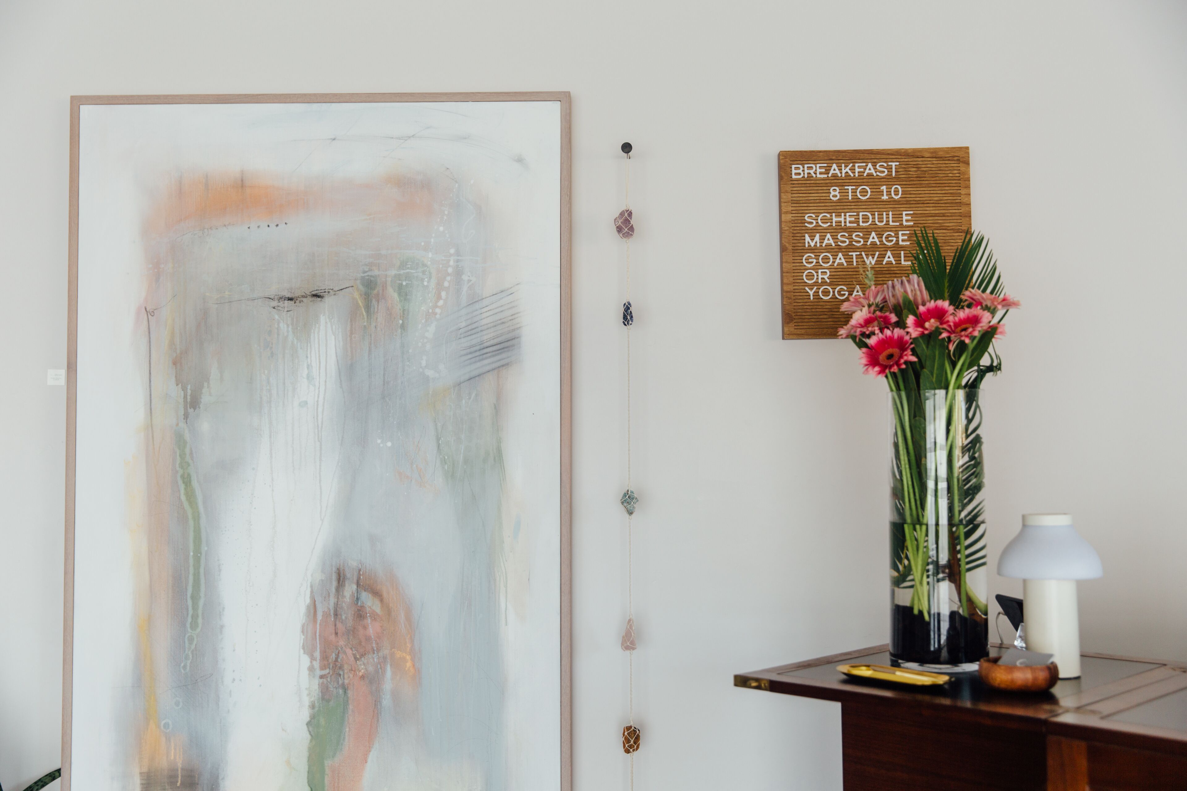 A room with a large abstract painting, a wooden sign with breakfast schedule, a vase with flowers, and a desk with a lamp.