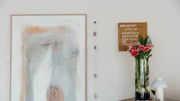 A room with a large abstract painting, a wooden sign with breakfast schedule, a vase with flowers, and a desk with a lamp.