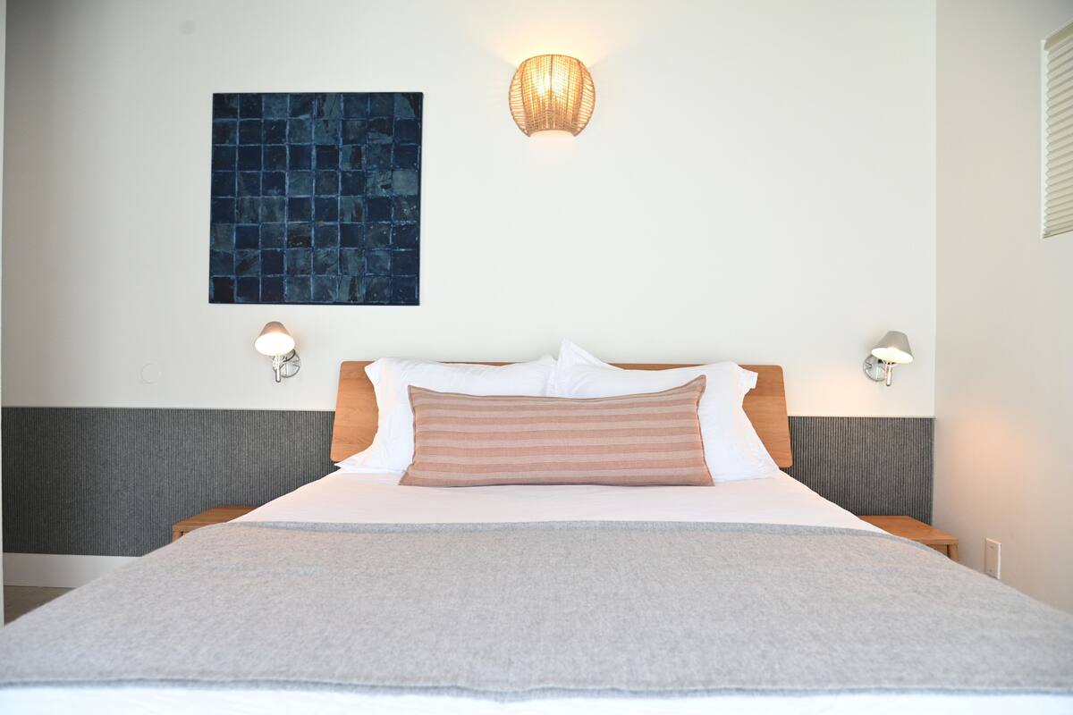 A hotel room with a bed, two bedside lamps, a wall-mounted light fixture, and a blue textured artwork.