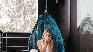 Une femme est assise dans un fauteuil suspendu de couleur turquoise sur une terrasse en bois, avec vue sur des arbres et un bâtiment en arrière-plan.