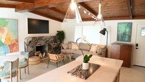Un salon moderne doté d’une cheminée, de poutres apparentes en bois au plafond, d’un canapé et d’un coin repas avec des chaises à motifs à carreaux.